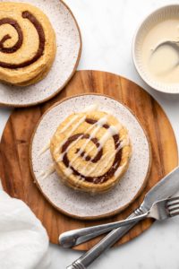 Two stacks of cinnamon roll pancakes on small white plates on round wood cutting board on marble background