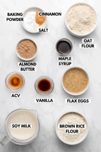 Ingredients for cinnamon roll pancakes in small white bowls on marble background. Clockwise text labels read oat flour, maple syrup, flax eggs, brown rice flour, soy milk, acv, vanilla, almond butter, baking powder, cinnamon, and salt