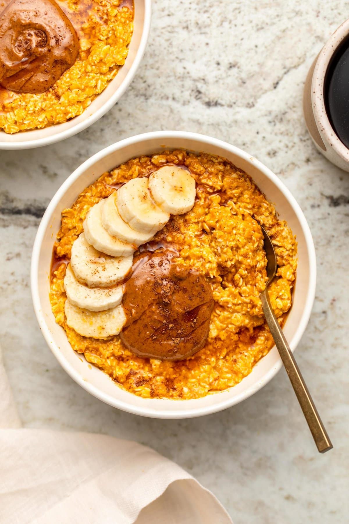 Pumpkin spice latte overnight oats in white bowl topped with banana, almond butter, and maple syrup