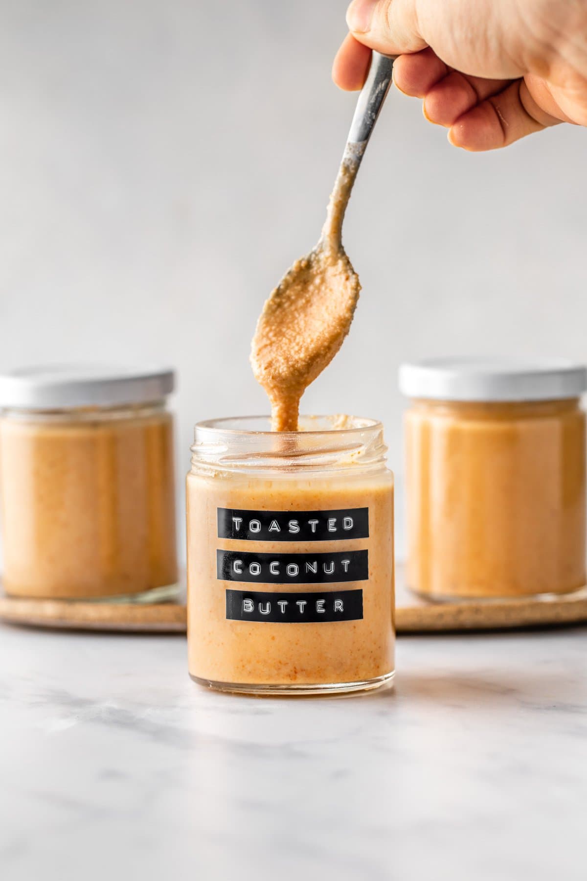 spoon drizzling toasted coconut butter into labeled jar on marble background