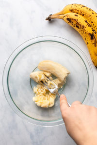 hand mashing banana in glass bowl on marble background