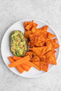 avocado half filled with guacasalsa alongside chips and carrots