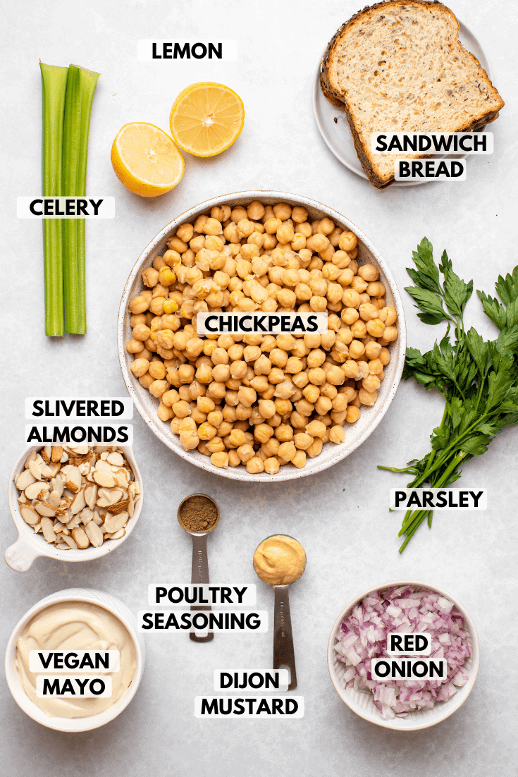 Ingredients for chickpea salad in small bowls on marble background. Clockwise text labels read sandwich bread, chickpeas, parsley, red onion, dijon mustard, poultry seasoning, vegan mayo, slivered almonds, celery, and lemon