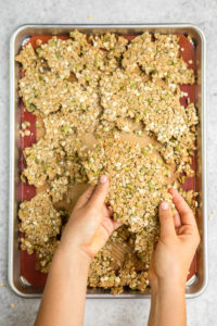 hands holding large cluster of tahini cardamom granola over baking tray