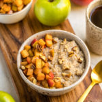 bowl of cinnamon apple oatmeal surrounded by apples on wood cutting board