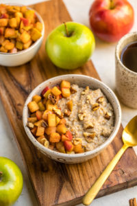 bowl of cinnamon apple oatmeal surrounded by apples on wood cutting board