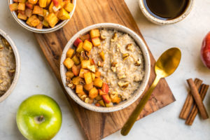 bowl of apple cinnamon oatmeal topped with cinnamon apples and walnuts on wood cutting board