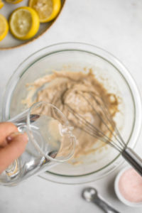 hand pouring water into glass bowl with thick tahini dressing