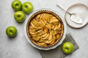 salted caramel apple tart on white pie stand surrounded by green apples and plates