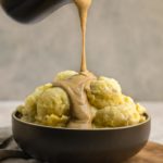 dark brown bowl of mashed potatoes with tahini gravy being poured over the top