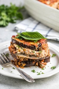 cross photo of layers of eggplant lasagna on small white plate topped with fresh basil leaf