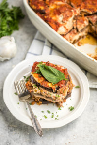 slice of vegan eggplant lasagna on small white plate with larger casserole dish in the background