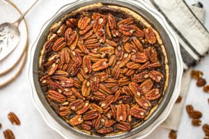 vegan pecan pie in silver pie tin on white marble background surrounded by pecan pieces