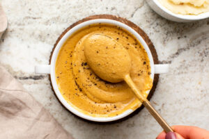Small white dish of vegan tahini gravy topped with black pepper and a gold serving spoon