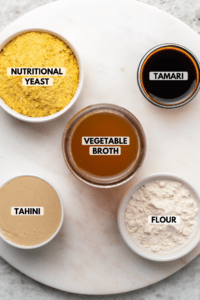Ingredients for Tahini Gravy in small bowls on marble countertop. Clockwise text labels read tamari, flour, tahini, vegetable broth, and nutritional yeast