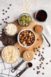 ingredients for chocolate hazelnut granola in small cups and bowls on round wooden cutting board with striped blue and white towel