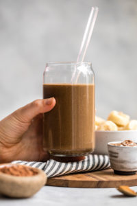 hand holding chocolate hemp smoothie in glass with straw over wooden cutting board