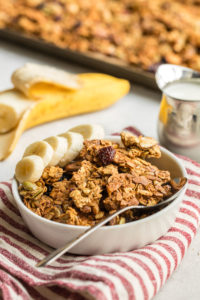 angled shot of white bowl filled with granola with sliced banana on the side on red striped napkin