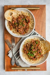 two bowls of lentil bolognese over pasta with toasted bread on white cutting board