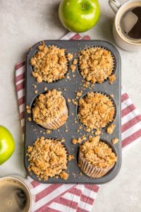 tray of baked apple cinnamon muffins with cups of coffee and fresh apples on light gray background