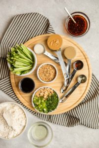 ingredients for spicy sesame noodles in measuring cups on round wood cutting board with black striped towel