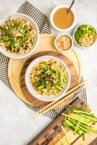 two white bowls of cooked sesame noodles topped with cucumber, green onion, chili oil, and peanuts