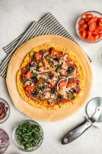 cooked sweet potato pizza crust surrounded by pizza cutter and toppings on white background