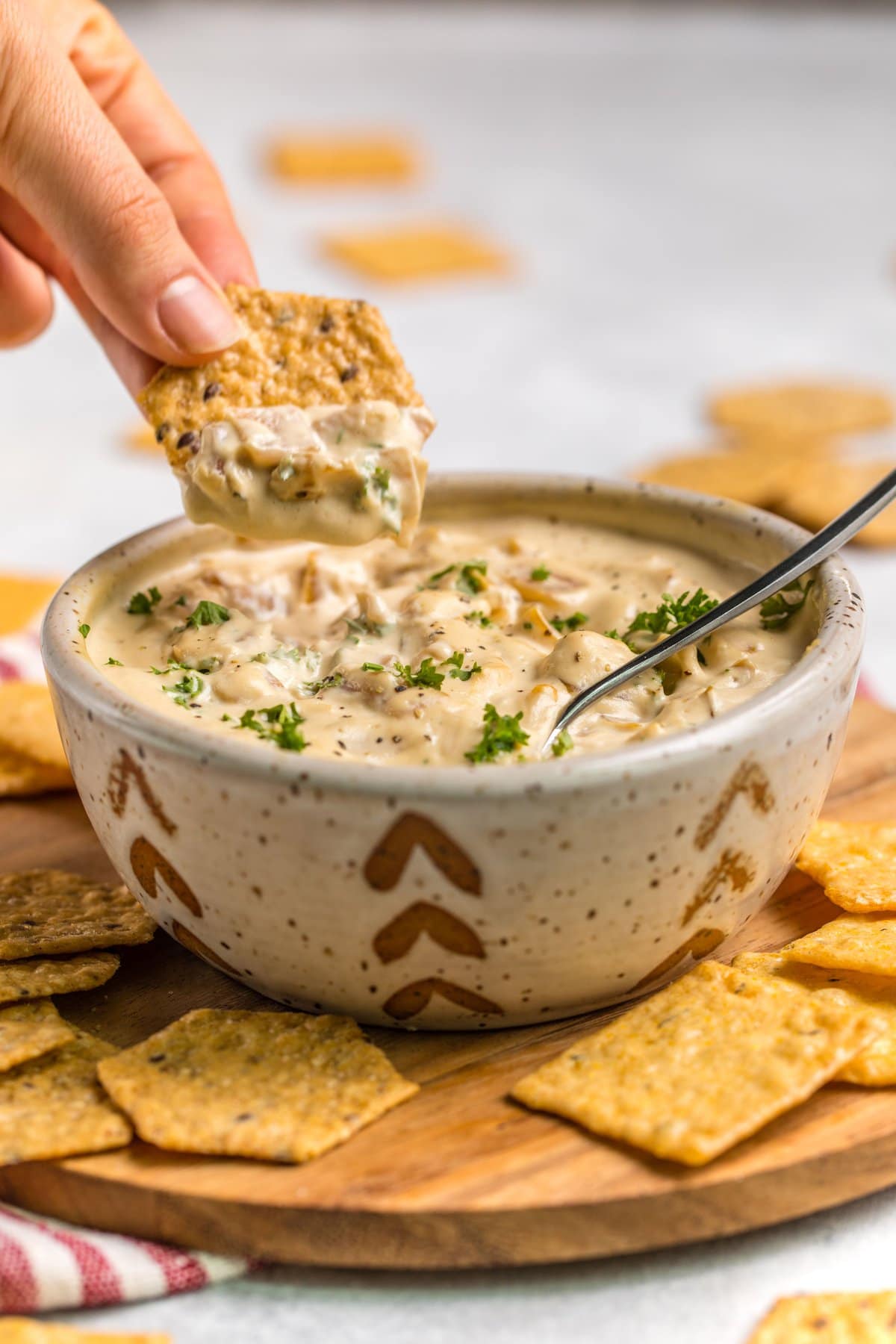 hand scooping cracker into vegan french onion dip in white speckled bowl on wood serving tray