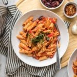 penne puttanesca topped with fresh basil and red pepper in white pasta bowl with black striped serving towel