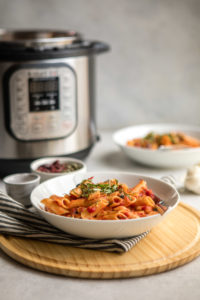 white pasta bowl of penne puttannesca on wood serving platter with instant pot in background