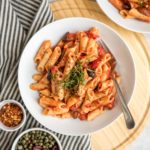 penne puttanesca topped with fresh basil and pepper in white pasta bowl on round wood cutting board