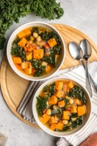 two white bowls filled with sweet potato kale soup on round wood serving tray with metal spoons