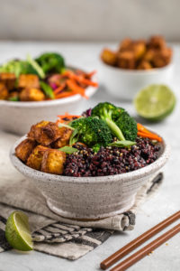 side view of two white speckled bowls filled with black rice, crispy tofu, broccoli, and asian slaw