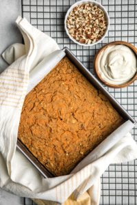 cooked healthy carrot cake on cooling rack with small bowls of frosting and chopped pecans