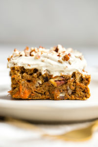 slice of healthy carrot cake topped with cashew frosting and chopped pecans on white plate