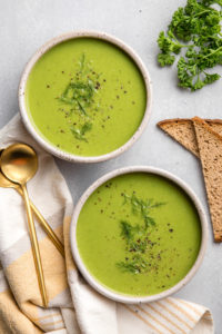 two bowls of green pea soup topped with fresh dill and black pepper with gold spoons