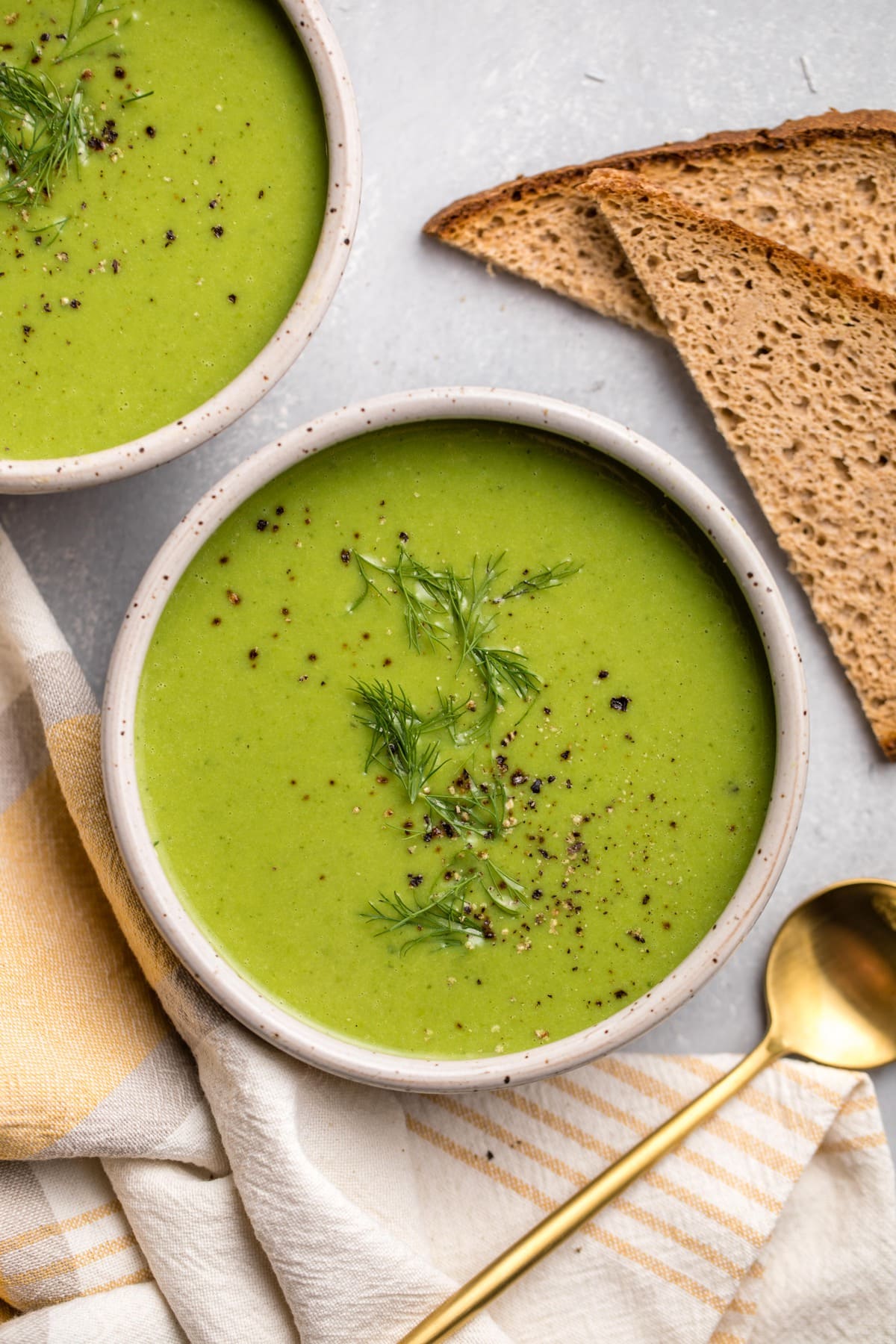 bowl of green pea soup with toast and golden spoon on grey textured background
