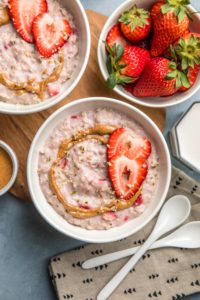 bowls of strawberry and cream oatmeal on round wooden cutting board topped with fresh strawberries