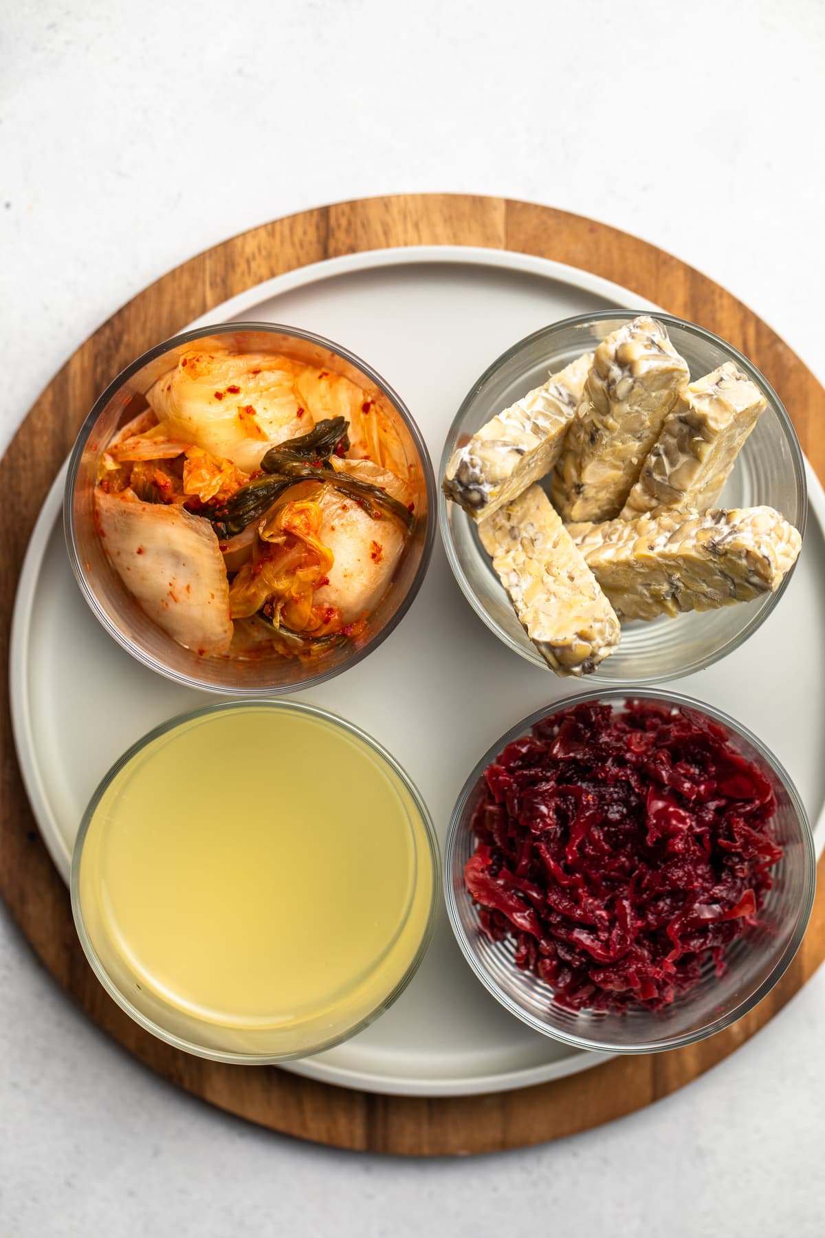 glass containers of kimchi, tempeh, kraut, and kombucha on round wooden cutting board