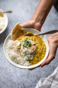 two hands holding bowl of dal tadka with rice, naan, and raita