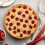 strawberry rhubarb pie with bowl of fresh strawberries and rhubarb on grey background