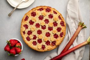 strawberry rhubarb pie with bowl of fresh strawberries and rhubarb on grey background