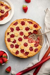 strawberry rhubarb pie with pie slice taken out of it on gray background with bowl of strawberries