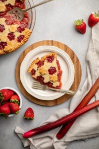 slice of strawberry rhubarb pie on white plate with pie in background and bowl of fresh strawberries
