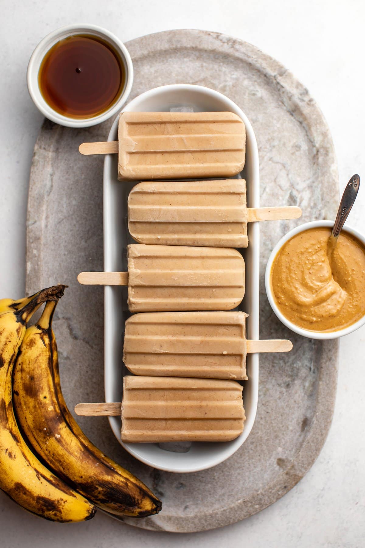 white tray of peanut butter banana popsicles with bowls of peanut butter and maple syrup and ripe bananas