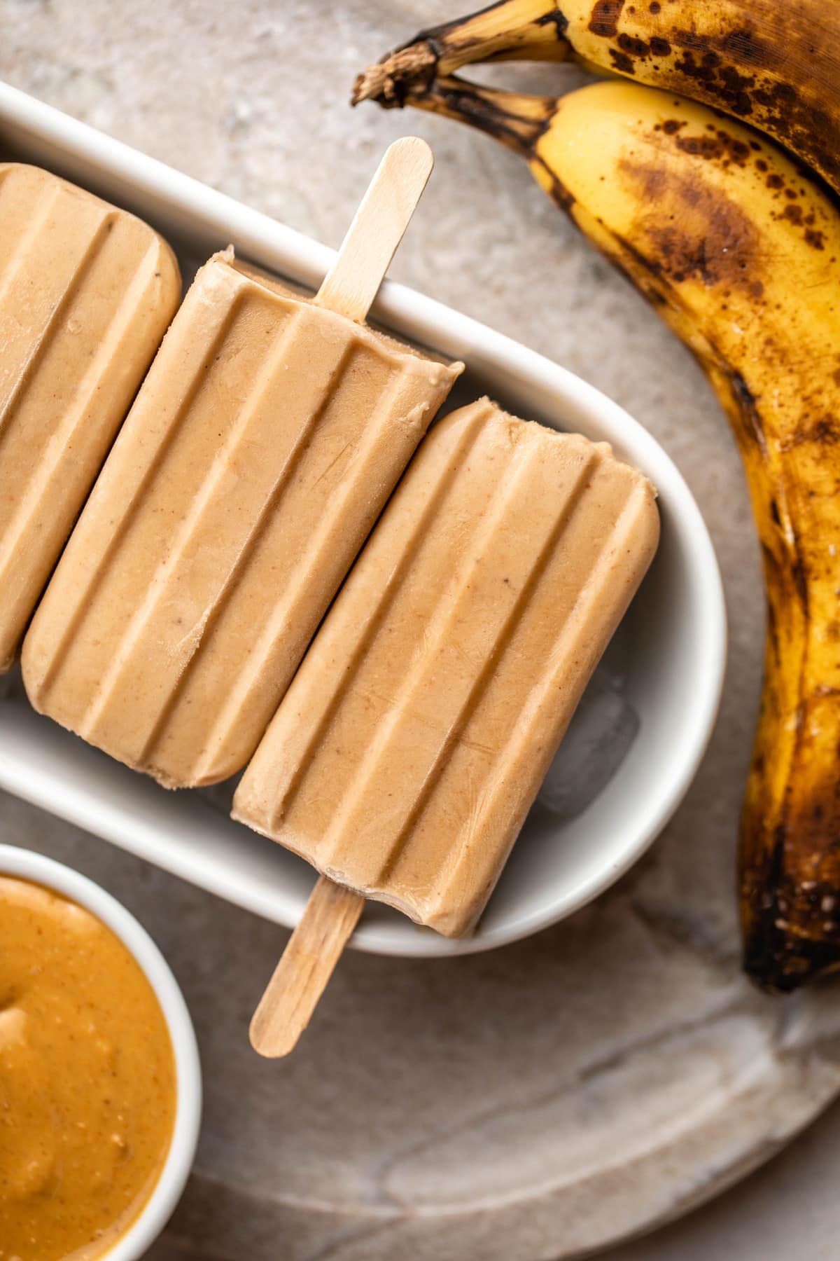 3 peanut butter popsicles with ripe bananas and bowl of peanut butter