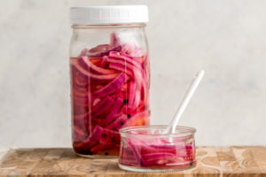 large mason jar and small glass dish of pickled red onions on wood cutting board