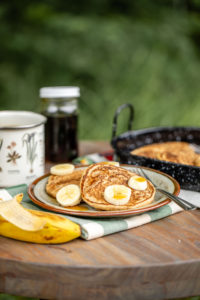 vegan pancakes topped with banana and maple syrup with mug of coffee on picnic table