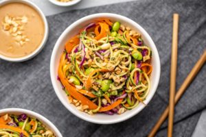 bowl of zucchini noodle salad with chopsticks and peanut sauce on dark linen