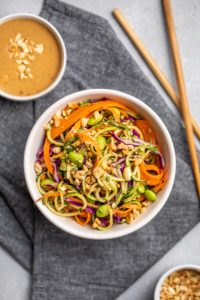 single bowl of zucchini noodle salad with bowls of peanut sauce and chopped peanuts